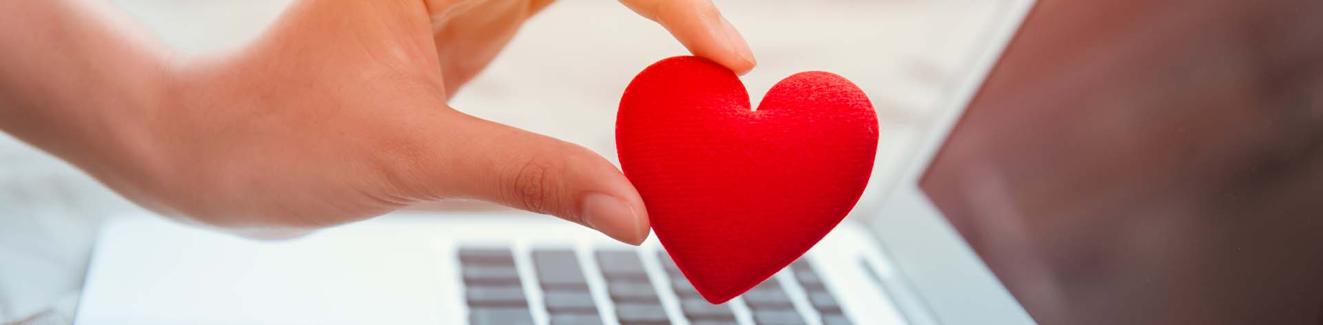 A hand holds a fabric heart; a laptop can be seen in the background.
